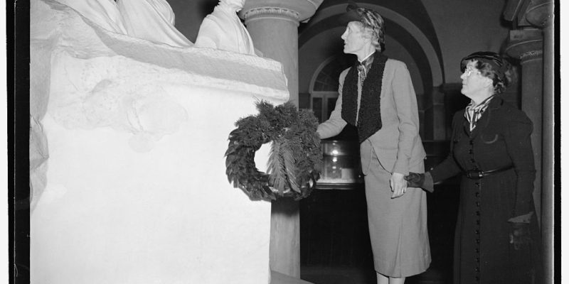 Two women laying a wreath in front of a statue composed of 3 busts of female suffragists
