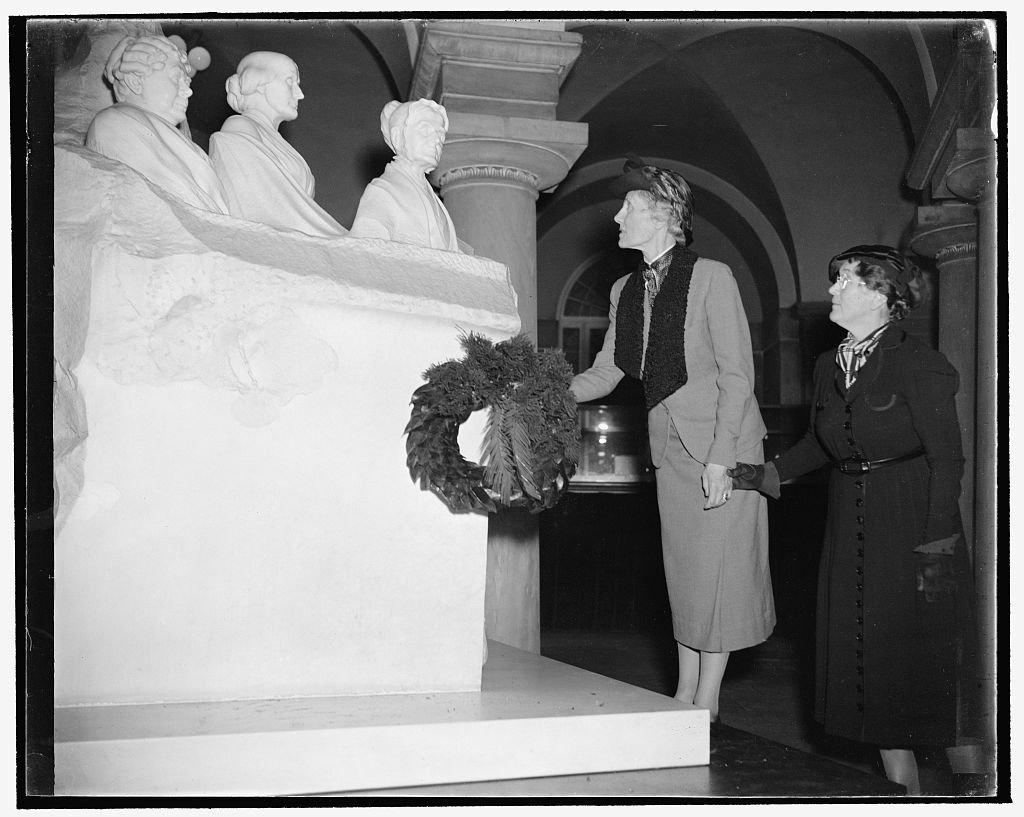 Two women laying a wreath in front of a statue composed of 3 busts of female suffragists